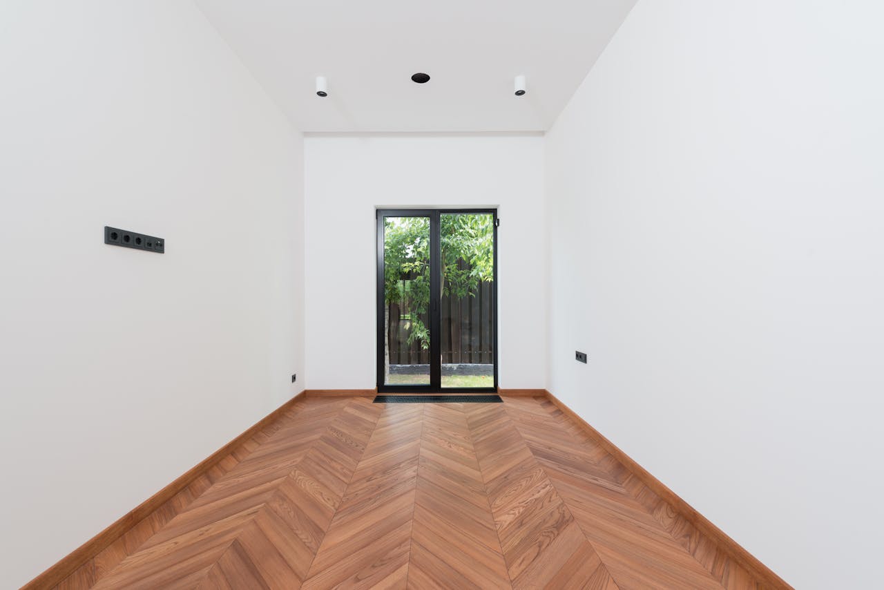 A clean, minimalist interior room with white walls, wooden parquet flooring, and a glass door.